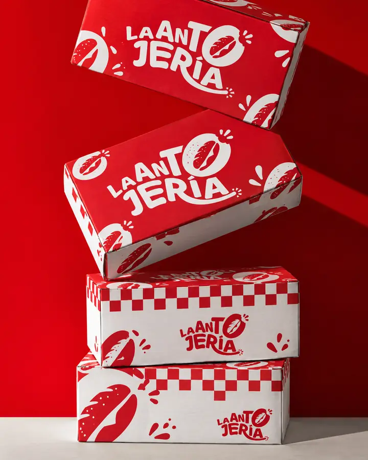 Cajas para llevar de La Antojería apiladas, en rojo y blanco con patrón de cuadros